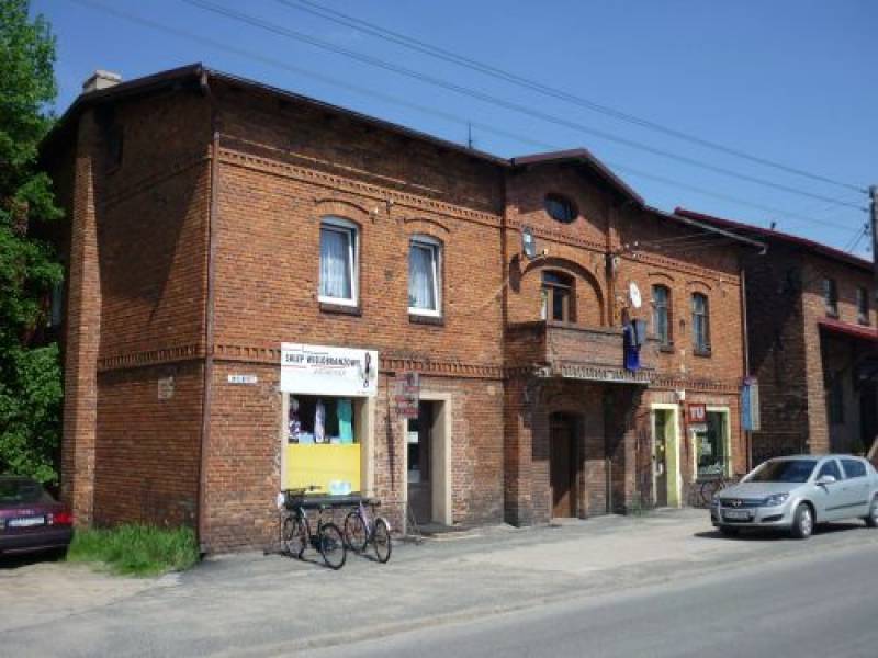 Budynek mieszkalny w Boronowie nr 1 (Frontowa część budynku mieszkalnego. Budynek w kolorze cegieł z białymi oknami i balkonem. Obok drzwi - witryna sklepowa. Przed budynkiem - stojak na rowery oraz zaparkowany samochód.)