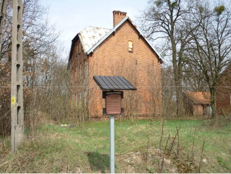 Zespół Budynków Szkolnych w Boronowie nr 2 (Boczna część tzw. „Wysokiej szkoły”. Budynek w kolorze cegieł z 1 niewielkim oknem. Na pierwszym planie - tabliczka informacyjna. W tle - drzewa oraz inne budynki.)