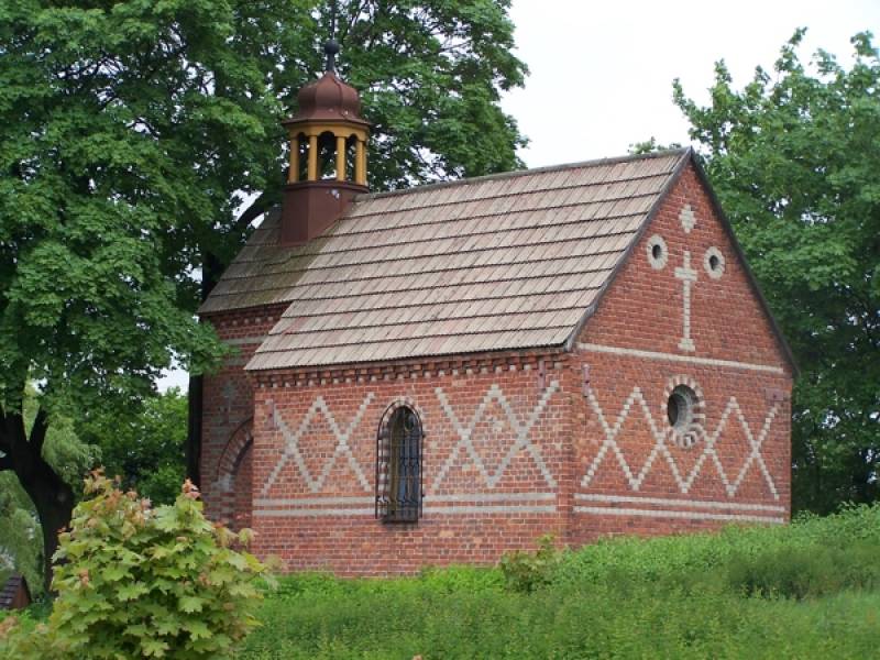 Kaplica Góra Oliwna w Woźnikach nr 1 (Boczna i tylnia część Kaplicy. Budynek z cegieł z jaśniejszymi wzorami i krzyżem. W tle drzewa.)