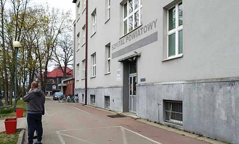 Budynek Szpitalny nr 1 (Szary budynek szpitala z ciemniejszym napisem: Szpital Powiatowy. Po lewej stronie widoczny mężczyzna i czerwone kosze na śmieci. W tle rowery, samochód oraz budynek.)