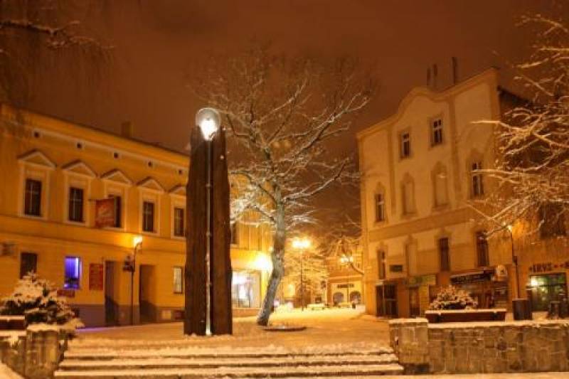 Pomnik Św. Teresy Benedykty od Krzyża - Edyty Stein nr 1 (Mały Rynek w Lublińcu nocą w zimie. Drewniany pomnik z aureolą. W tle drzewo oraz budynki.)