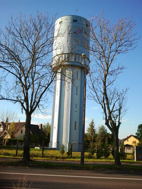 Wieża ciśnień (Wysoka wieża w kolorze niebieskim z elementami w kolorze białym. Liczne białe okna. Przed wierzą ulica oraz drzewa. W tle budynki oraz krzewy.)
