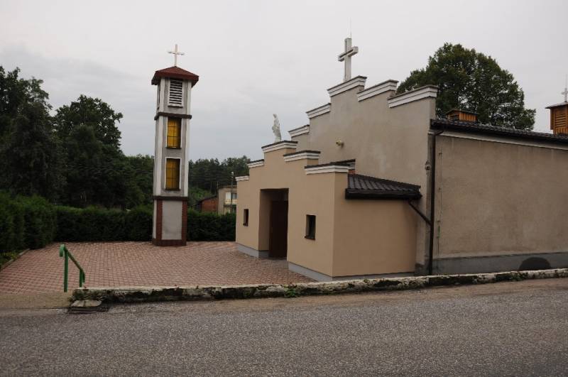 Parafia Najświętszej Maryi Panny Królowej w Kokotku nr 1 (Boczna część Kościoła. Jasny budynek z brązowymi drzwiami i dwoma małymi oknami. Budynek zakończony białym krzyżem. Z przodu widoczna wolnostojąca wieża zakończona białym krzyżem. W tle krzewy, drzewa oraz budynek.)