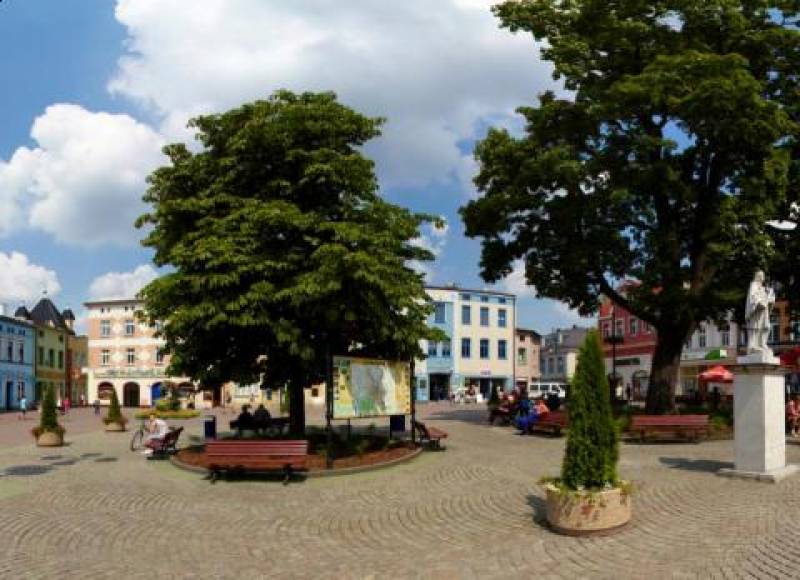 Rynek - Plac Konrada Mańki (Rynek w Lublińcu. Brązowe ławki ustawione wokół dwóch drzew. Przy jednej z nich widoczna tablica z planem miasta. Z prawej biały pomnik. W tle budynki)