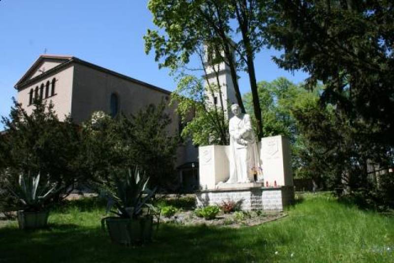 Pomnik Św. Eugeniusza de Mazenod nr 1 (Pomnik Św. Eugeniusza de Mazenod w kolorze białym na podwyższeniu. Pomnik otoczony krzewami i drzewami. W tle widoczny fragment budynku.)