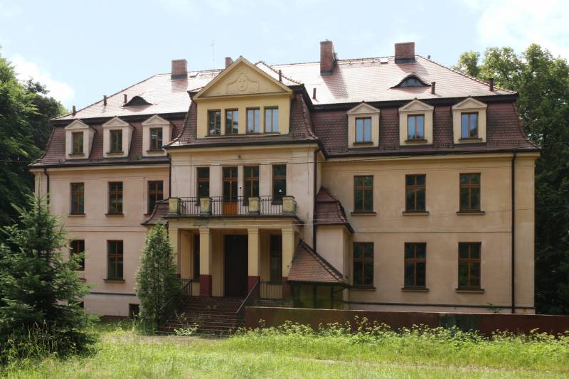 Zespół Pałacowo-Parkowy w Ciasnej nr 1 (Pałac od strony wejścia głównego, wokół trawa i las, budynek dwu piętrowy z dużą ilością okien oraz balkonem)